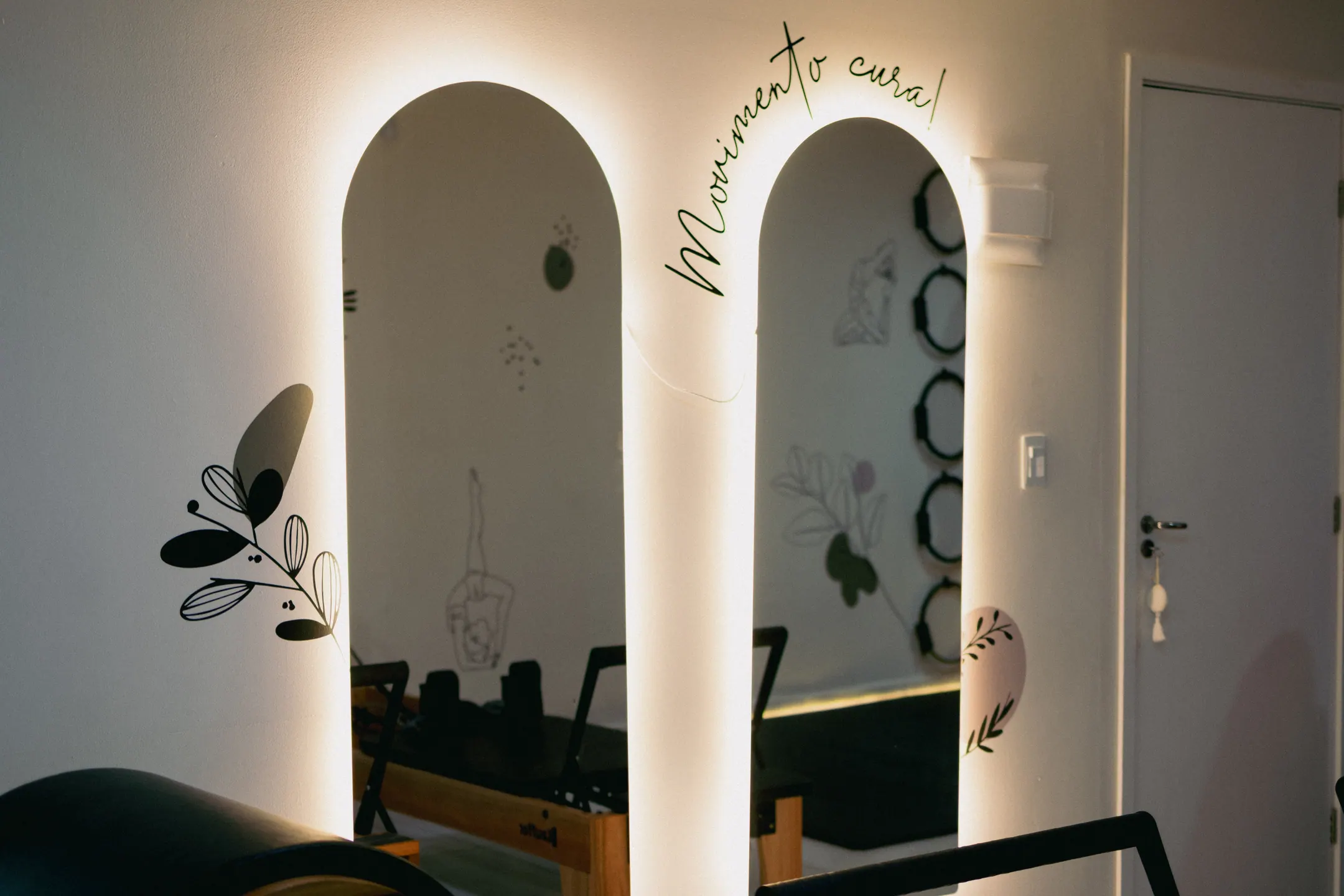 Dois vãos em arco iluminados por trás na parede branca do estúdio, com a frase Movimento cura escrita em cursiva ao lado