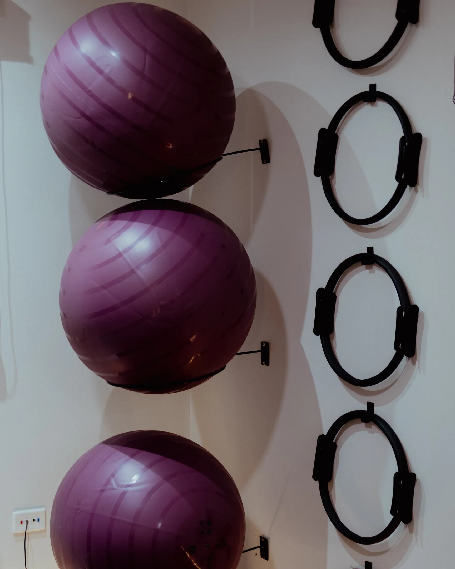 Três bolas de pilates roxas encaixadas em suportes na parede, ao lado de uma coluna de arcos flexíveis pretos