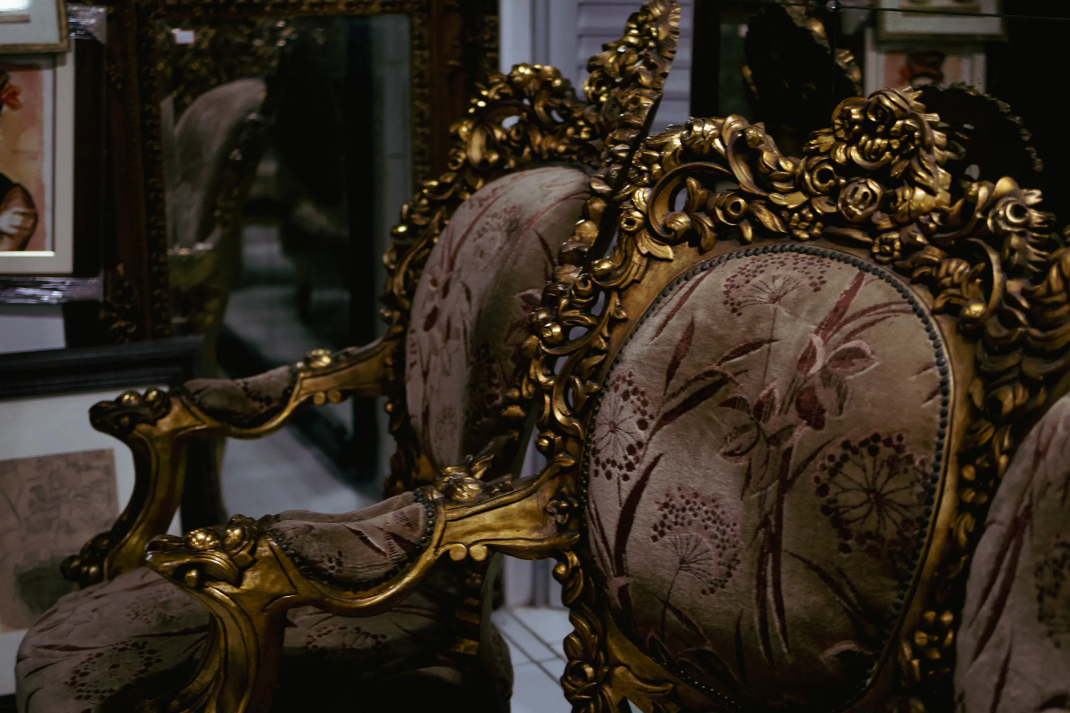 Poltrona dourada de espaldar entalhado e estofado floral, refletida num espelho de moldura ornamentada