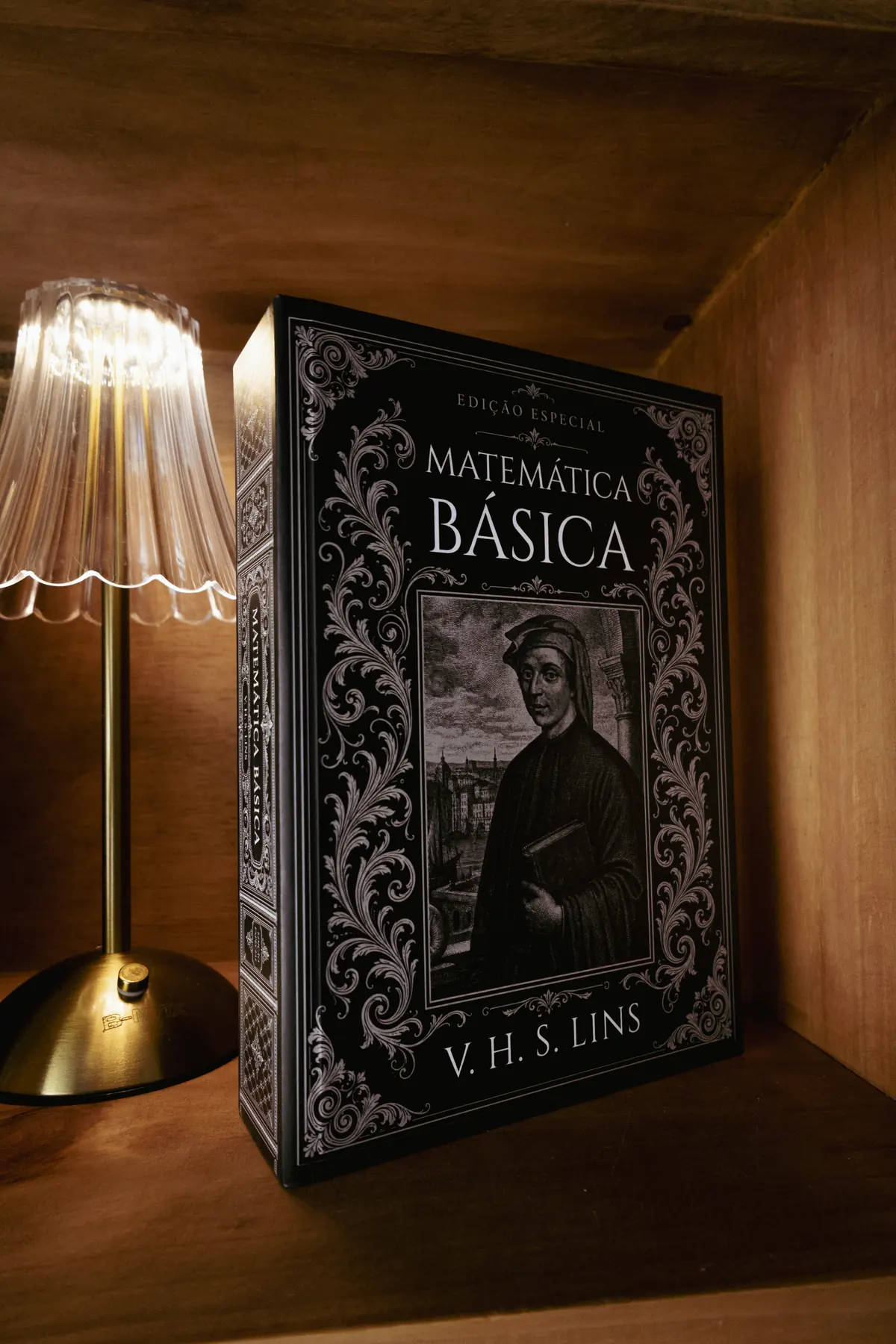 Livro apoiado em um nicho de madeira ao lado de um abajur de cúpula plissada aceso