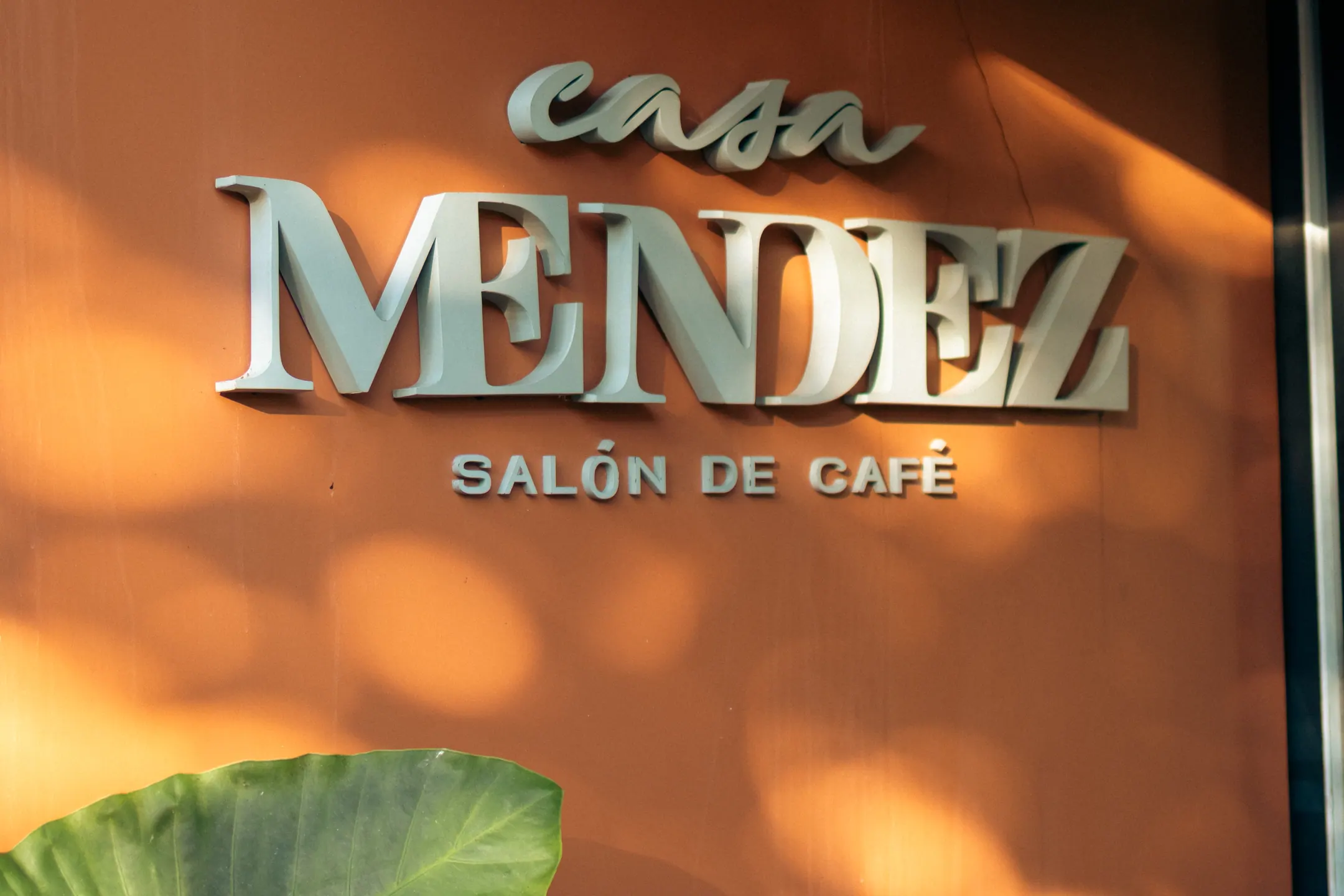 Letreiro em relevo com o nome Casa Mendez, salón de café, sobre a parede terracota, com a sombra de uma folha atravessando