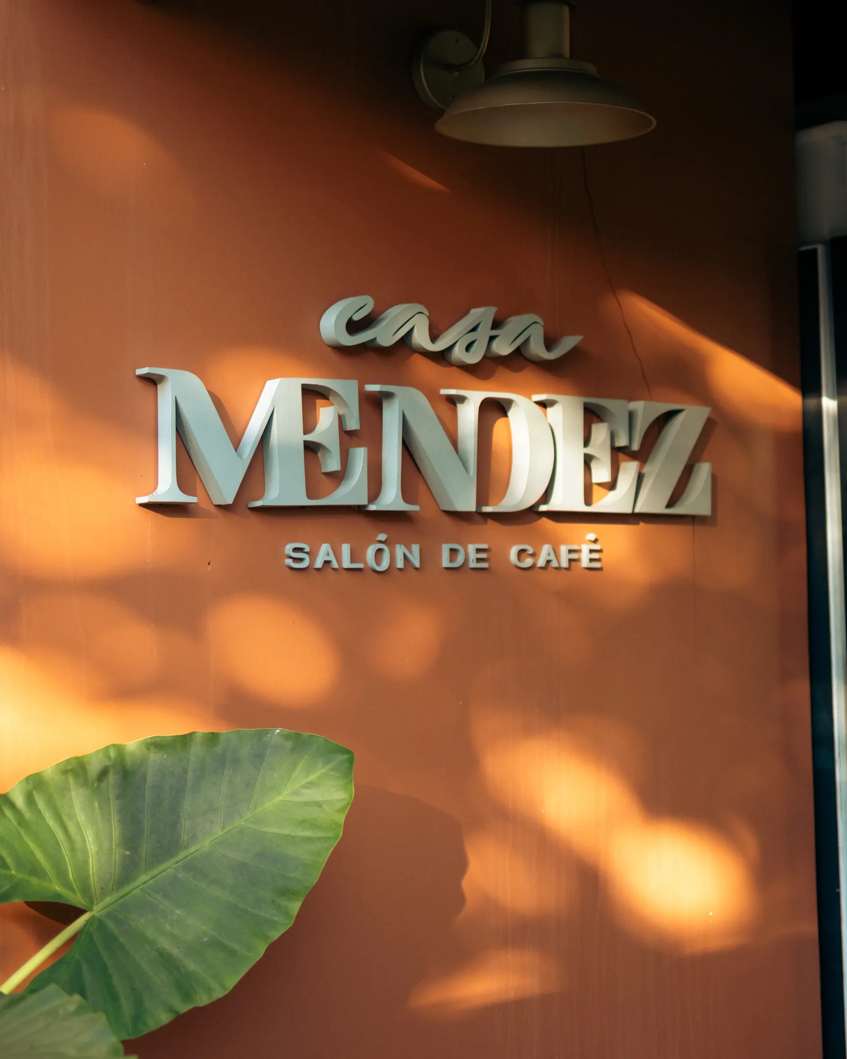 Letreiro em relevo com o nome Casa Mendez, salón de café, sobre a parede terracota, com a sombra de uma folha atravessando