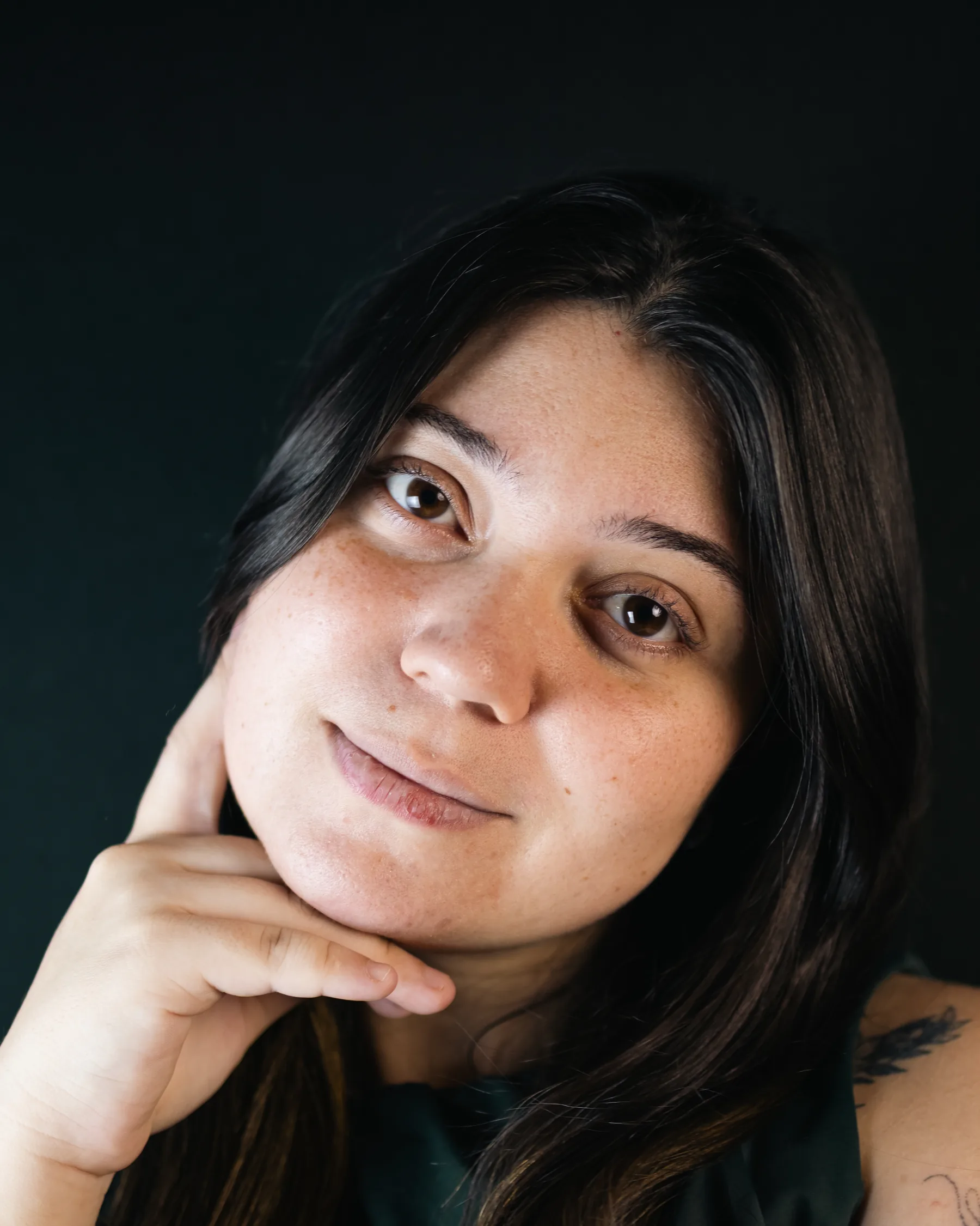 Retrato de estúdio em close: mulher de cabelo longo e vestido verde, mão apoiada no queixo, olhando para a câmera sobre fundo escuro