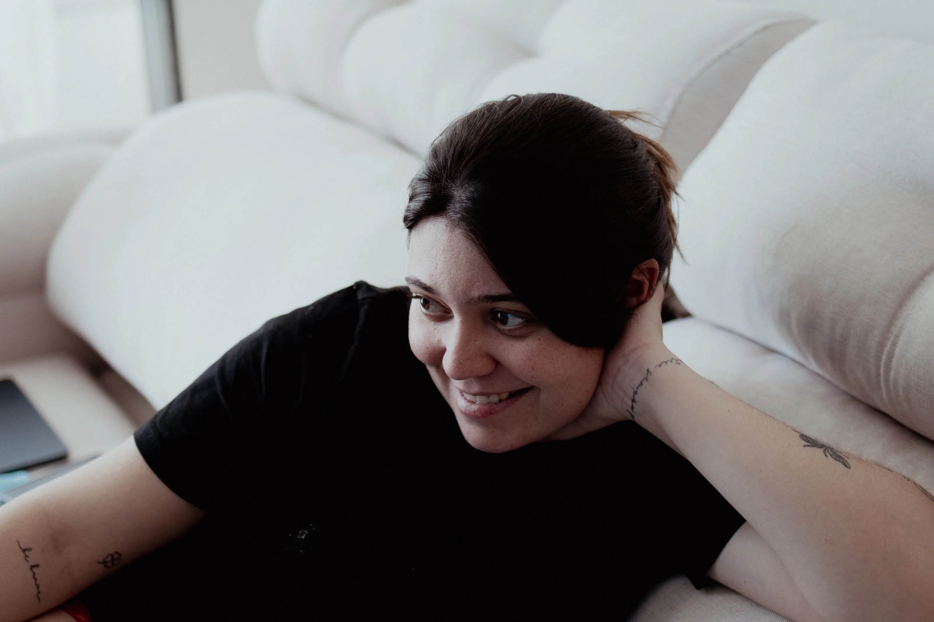 Mulher de camiseta preta deitada num sofá claro, apoiada no próprio braço, sorrindo e olhando para o lado em luz de janela