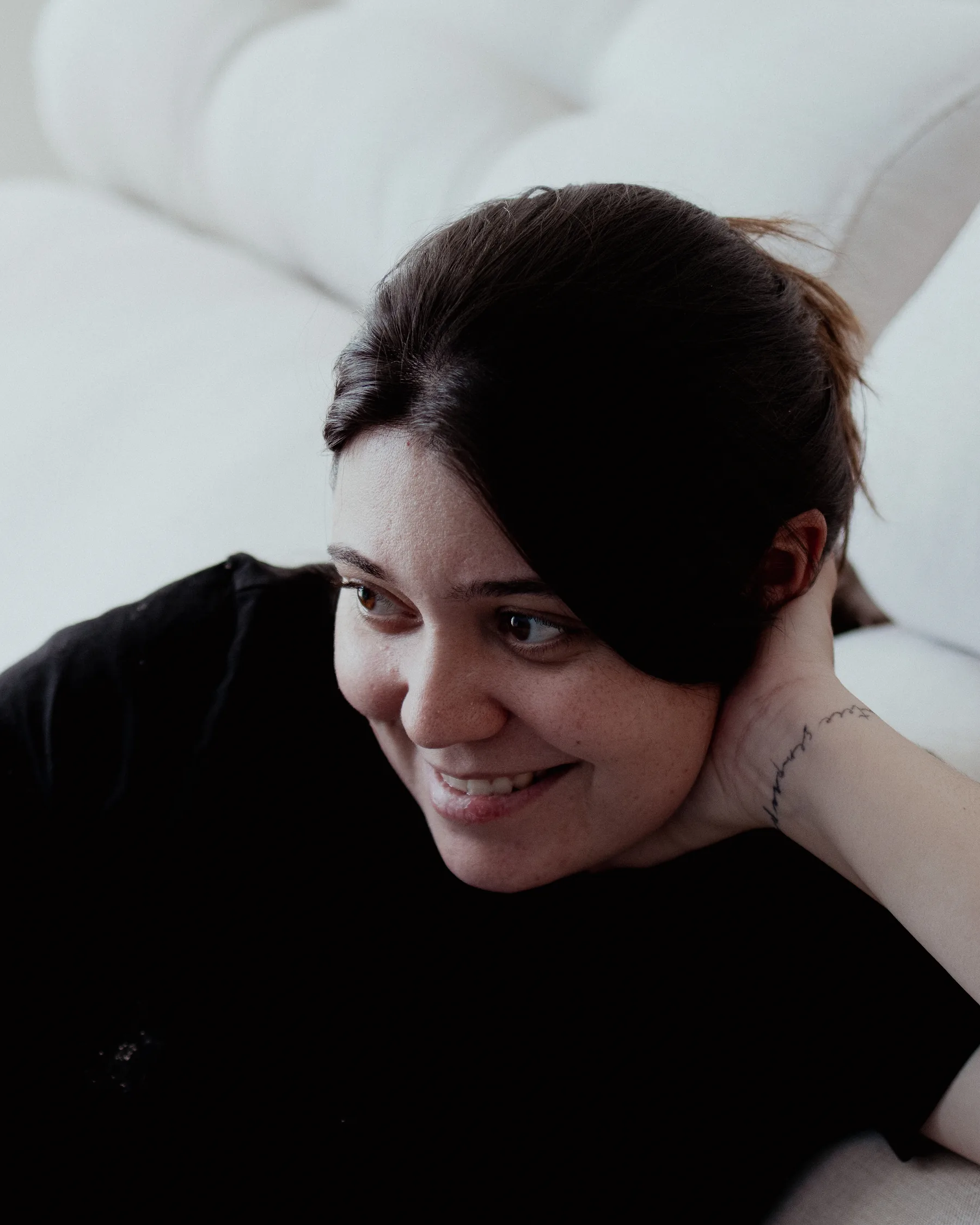 Mulher de camiseta preta deitada num sofá claro, apoiada no próprio braço, sorrindo e olhando para o lado em luz de janela