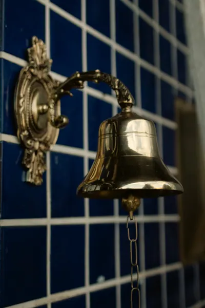 Sino de bronze com suporte ornamentado, preso na parede de azulejo azul