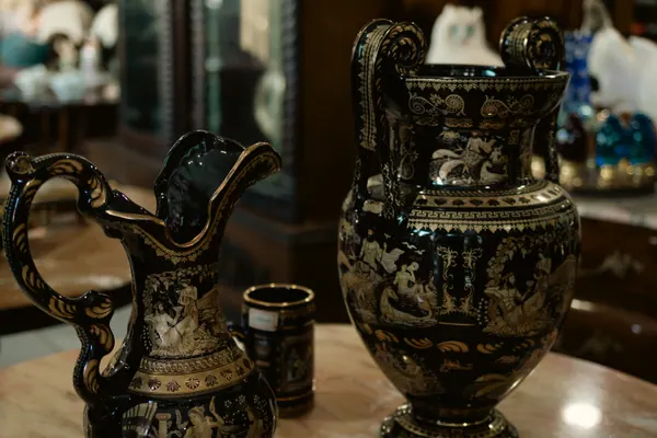 Jarra e vaso negros com cenas mitológicas douradas, sobre a mesa da loja