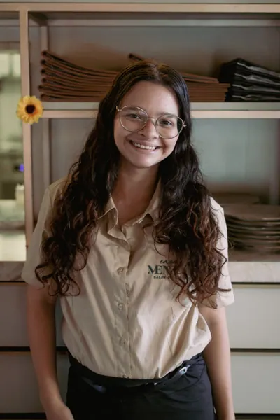 Atendente da Casa Mendez sorrindo para a câmera, de camisa bege com o nome da casa bordado, diante da prateleira de louça