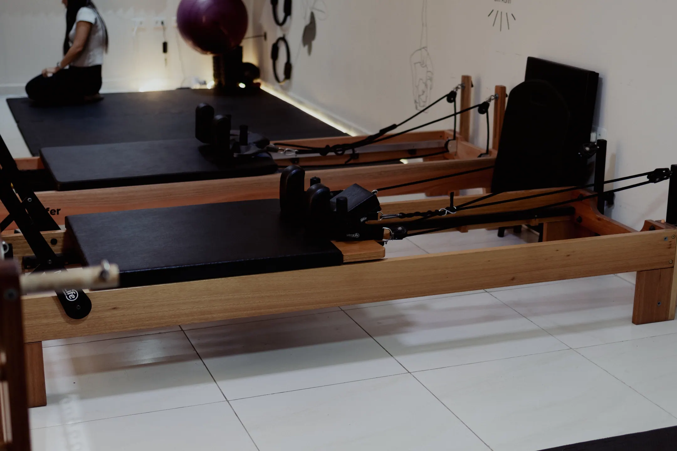 Dois reformers alinhados na sala clara, com uma aluna sentada de costas no colchonete ao fundo