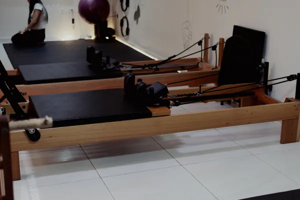 Dois reformers alinhados na sala clara, com uma aluna sentada de costas no colchonete ao fundo