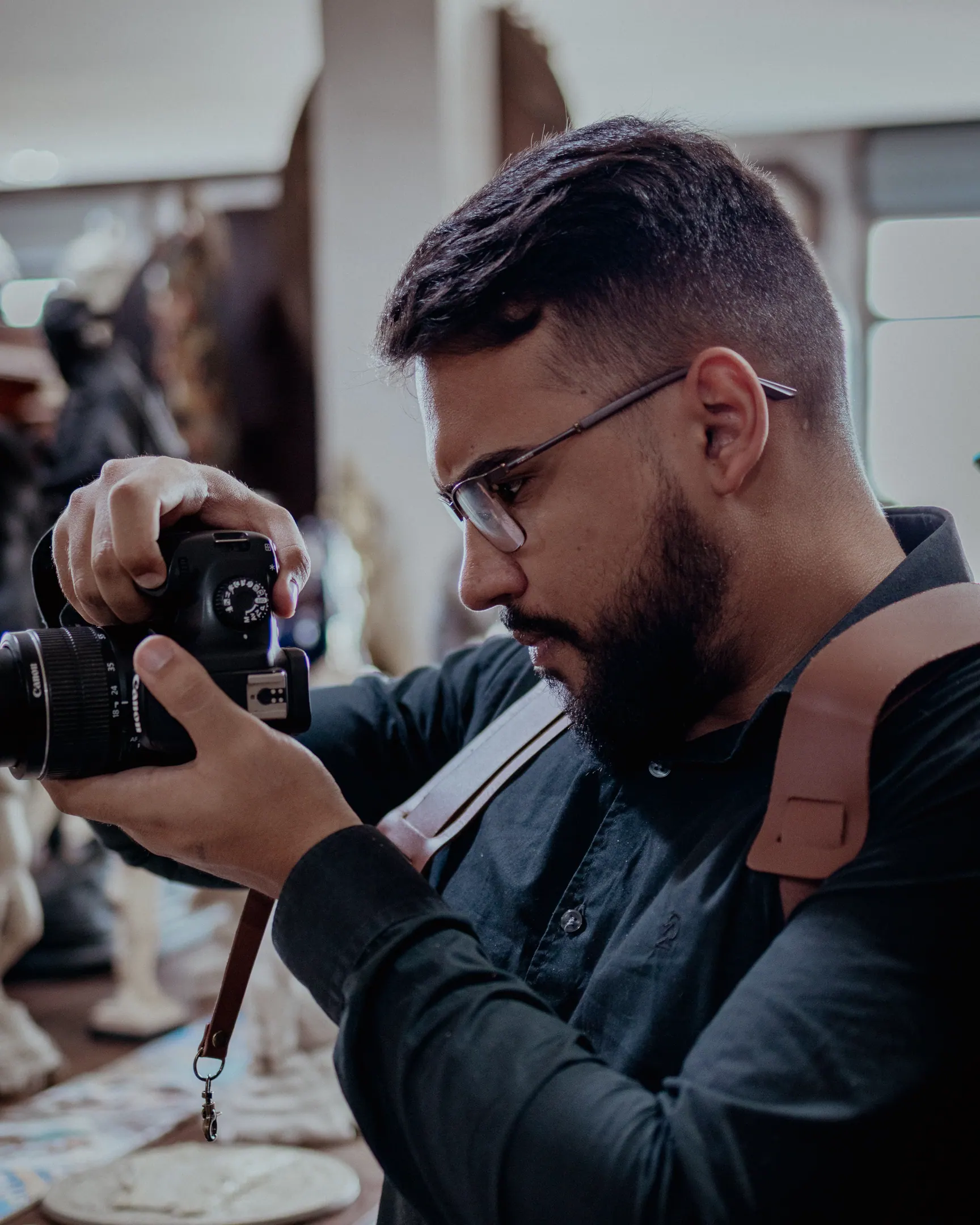 Victor Lins fotografando dentro do Antiquário São Bernardo, em Gravatá: de perfil, óculos, câmera erguida na mão, com as peças da loja desfocadas ao fundo