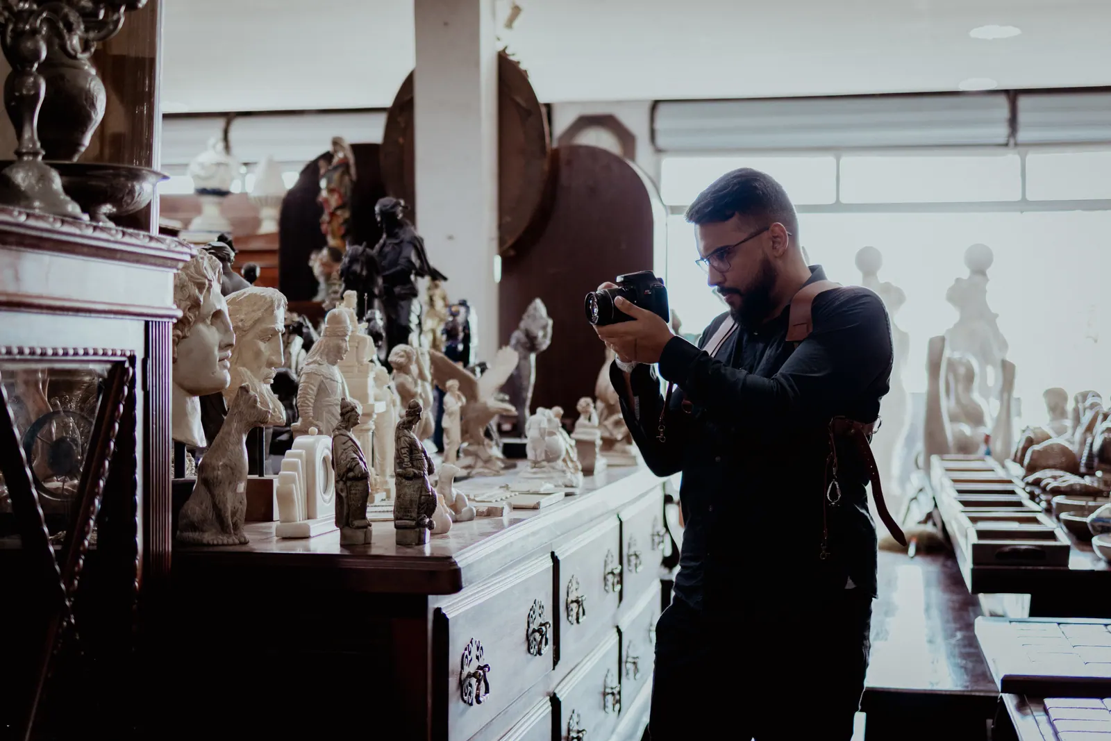 Victor Lins de pé no salão do Antiquário São Bernardo, câmera na altura do rosto, fotografando uma cômoda coberta de bustos e esculturas de mármore
