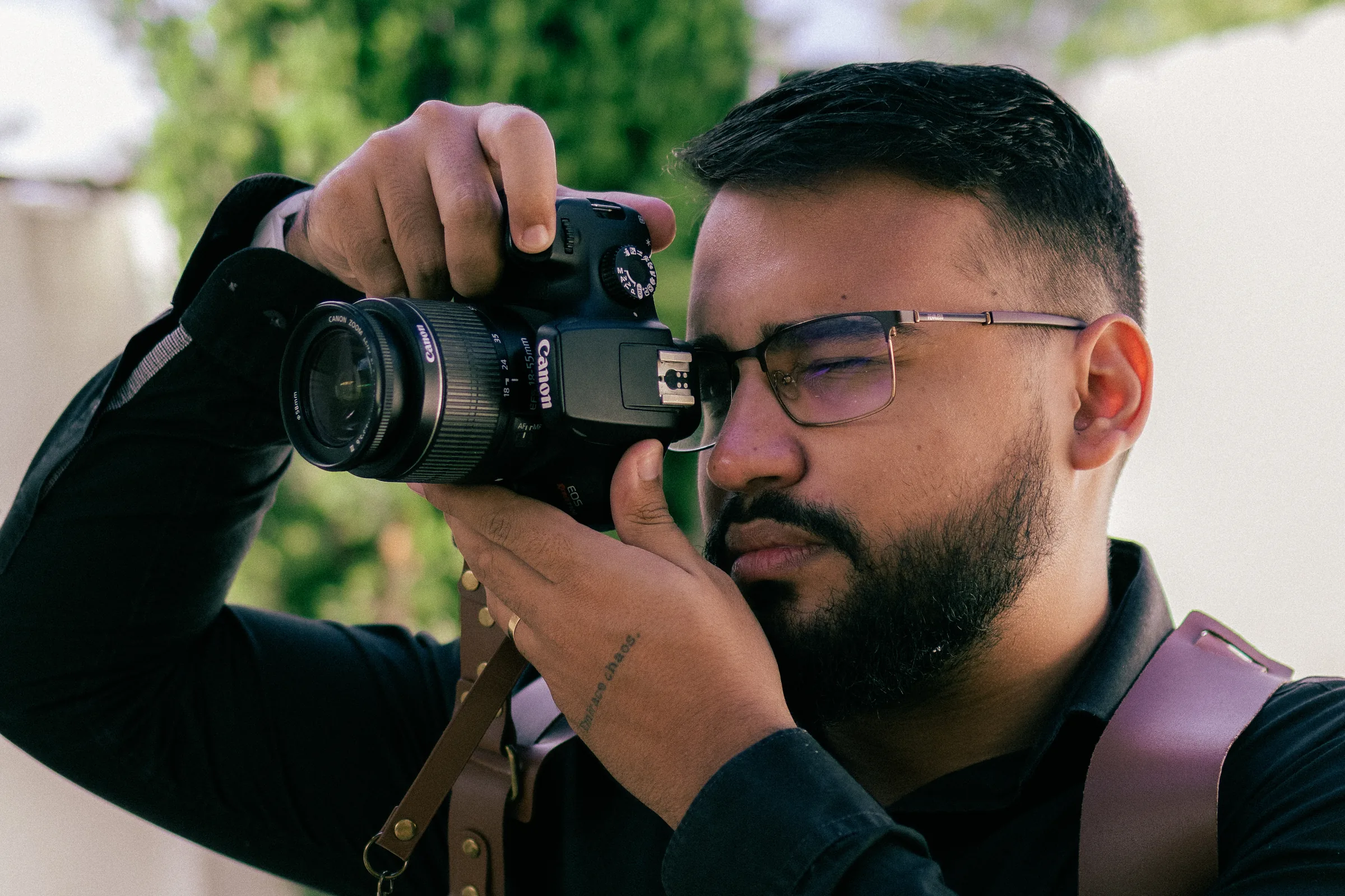Victor Lins fotografando com a câmera no olho, de camisa preta e suspensório de couro, em luz natural de fim de tarde